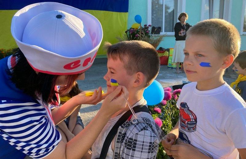 Знімок крупним планом, на якому дівчина-аніматор (у білому капелюсі з червоним написом та тільняшці) обережно наносить пензликом патріотичний аквагрим у кольорах українського прапора на щоку маленькому хлопчику в картатій сорочці. Поруч стоїть інший хлопчик із синім мазком на щоці.