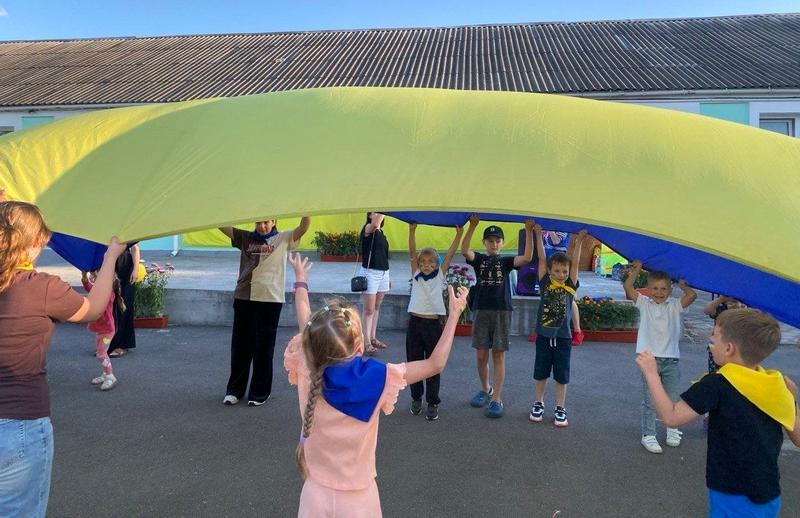 Велика група дітей та підлітків стоїть у колі на асфальтовому майданчику, піднімаючи та тримаючи над головами величезне полотно ігрового парашута синьо-жовтих кольорів (на вигляд як прапор України). Поруч стоїть дорослий, спостерігаючи за грою.