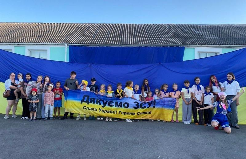 Повноформатне фото великої групи дітей та дорослих, які стоять на асфальті на фоні величезного синьо-жовтого прапора України, розтягнутого на всю стіну будівлі. Спереду вони тримають банер з написом 'Дякуємо ЗСУ! Слава Україні! Героям Слава!'. Справа — аніматор у костюмі моряка.