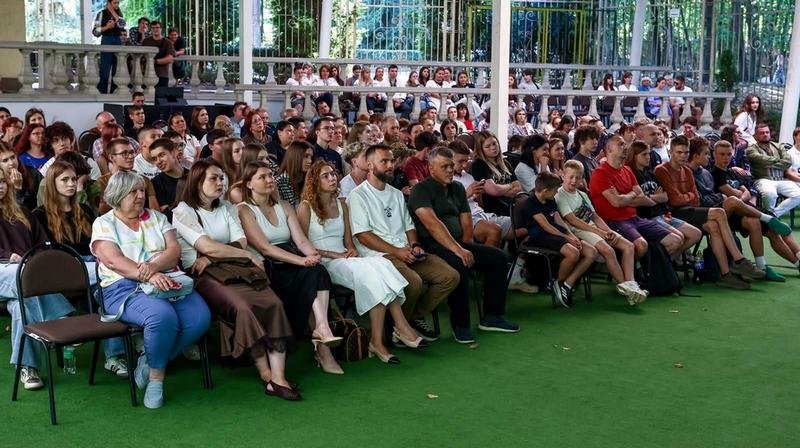 Глядачі різного віку, серед яких молодь, ветерани та представники громади, сидять у рядах літнього кінотеатру та уважно спостерігають за показом фільму.