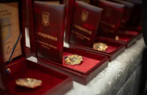 Ряд розкритих нагородних футлярів із темно-червоного оксамиту, що стоять на столі. У кожному футлярі знаходиться золотистий нагрудний знак «Мати-героїня» та посвідчення до нього з написом «Посвідчення до почесного звання України».