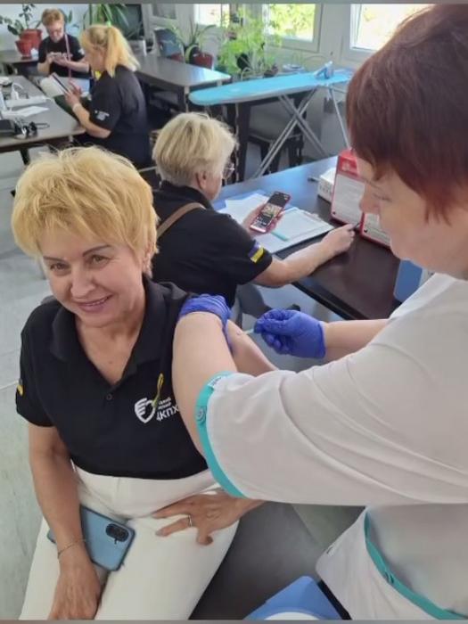 Медична сестра в рукавичках робить укол у плече усміхненій жінці в чорній футболці з логотипом Центру контролю та профілактики хвороб (ЦКПХ). На задньому плані видно інших учасників заходу в приміщенні.