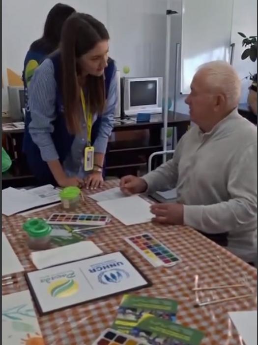 Представниця благодійного фонду спілкується з чоловіком похилого віку під час заходу. На столі розкладені акварельні фарби, папір та інформаційні матеріали з логотипами БФ «Рокада» та УВКБ ООН (UNHCR).