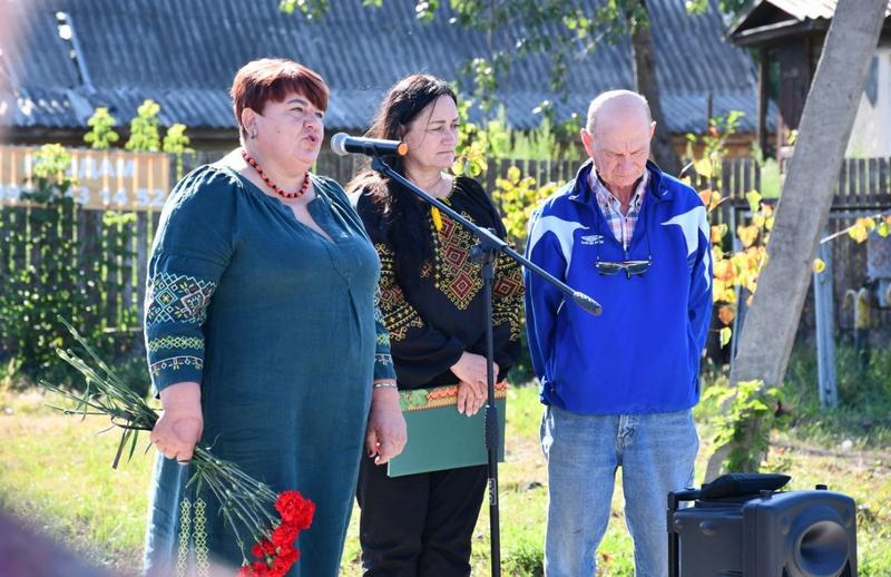 Заступник Макарівського селищного голови Марина Радченко у зеленій сукні з вишивкою виступає зі сценарною мовою під час відкриття Алеї пам'яті. Поруч із нею біля мікрофона стоять ведуча у вишиванці та чоловік у синій куртці, які беруть участь у церемонії.