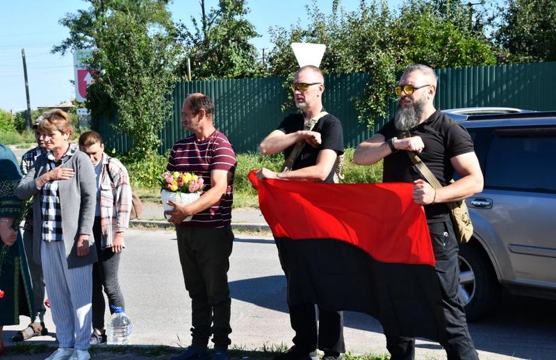 Два чоловіка у чорному одязі та тактичних окулярах тримають розгорнутий червоно-чорний прапор під час церемонії. Інші учасники заходу стоять поруч, тримаючи руки на серці під час звучання гімну.