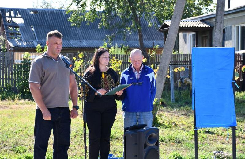 Офіційна частина відкриття меморіалу. Біля мікрофона виступає староста Калинівського старостинського округу Роман Ляшенко у футболці-поло. Поруч стоять жінка у вишиванці та чоловік у синій куртці. Праворуч — загорнутий у синю тканину пам'ятний стенд, готовий до відкриття.