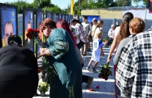 Люди стоять у черзі вздовж меморіальних стендів на Алеї пам'яті у Фасівочці, тримаючи в руках квіти для покладання. Серед присутніх є діти. На задньому плані видно державний прапор.