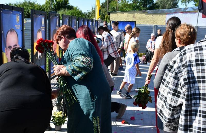 Люди стоять у черзі вздовж меморіальних стендів на Алеї пам'яті у Фасівочці, тримаючи в руках квіти для покладання. Серед присутніх є діти. На задньому плані видно державний прапор.