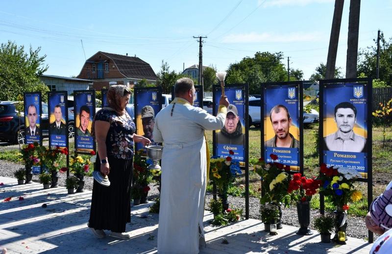 Священник у світлому облаченні освячує меморіальні стенди на Алеї пам'яті у Фасівочці. Жінка поруч тримає чашу. Позаду видно портрети захисників, зокрема Миколи Єфімова та Руслана Романова, прикрашені квітами.