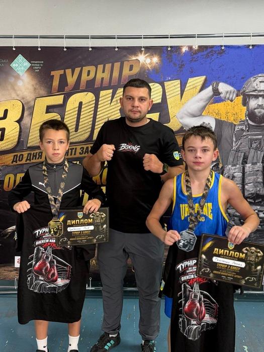 Тренер у чорній футболці стоїть по центру між двома юними боксерами на тлі банера турніру з боксу пам’яті Олексія Наконечного «Артиста». Спортсмени тримають дипломи, медалі за 1 та 2 місця та пам'ятні футболки.