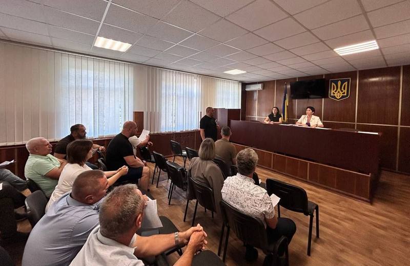Загальний вигляд зали засідань Макарівської селищної ради. Депутати сидять обличчям до президії, де за столом знаходяться головуюча на засіданні та секретар. На стіні за президією — Державний Герб України та прапор України. Керівник структурного підрозділу виступає з промовою.