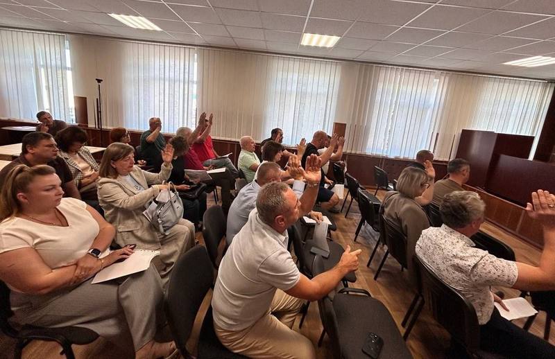 Вигляд збоку на залу засідань Макарівської селищної ради. Депутати сидять на стільцях та піднімають руки на знак підтримки рішення. Світло падає з великих вікон із жалюзі. Більшість присутніх зосереджені на голосуванні.