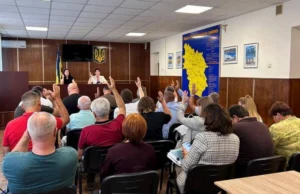 Депутати Макарівської селищної ради піднімають руки під час голосування. На задньому плані видно президію та велику мапу Макарівської громади на стіні. Засідання проходить у залі зі світлою стелею та дерев'яними панелями.