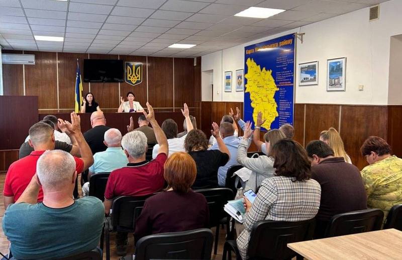 Депутати Макарівської селищної ради піднімають руки під час голосування. На задньому плані видно президію та велику мапу Макарівської громади на стіні. Засідання проходить у залі зі світлою стелею та дерев'яними панелями.