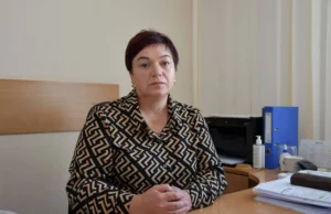 Портрет Валентини Курач, жінки середнього віку з короткою стрижкою, одягненої в блузку з геометричним візерунком. Вона сидить за робочим столом, склавши руки перед собою, на тлі офісного інтер'єру з папками та технікою.