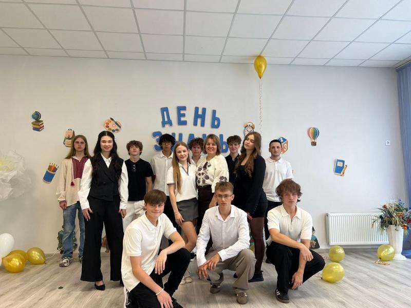 Старшокласники та вчителька у світлому класі з на написом «День знань» на стіні та жовтими повітряними кульками.