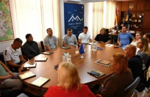 Офіційна зустріч у кабінеті Макарівського селищного голови з нагоди Дня фізичної культури і спорту. Учасники сидять навколо великого столу, на якому розкладені почесні нагороди для тренерів та вчителів. Позаду видно банер «Rebuild Makariv».