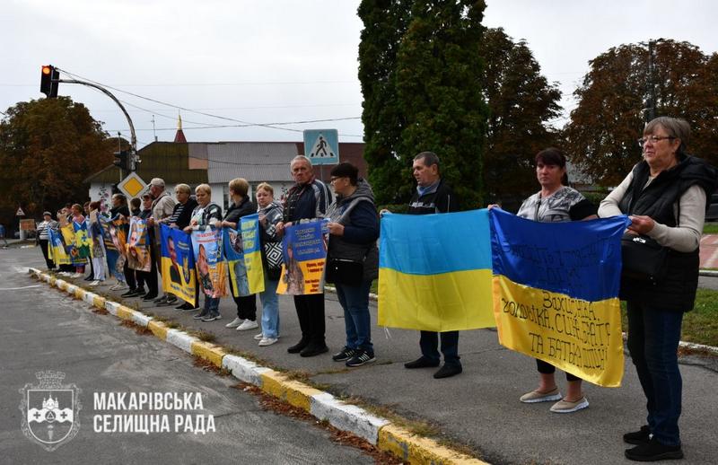 Учасники мирної акції в Макарові стоять уздовж дороги з синьо-жовтими прапорами та плакатами із зображеннями полонених і зниклих безвісти військових.