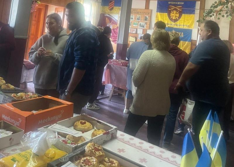 Люди спілкуються біля столів із домашніми смаколиками на благодійній ярмарку у приміщенні з українськими стягами