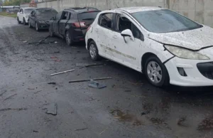 Чотири пошкоджені автомобілі стоять у ряд на дорозі. Кузов білої машини на передньому плані вкритий осколковими пошкодженнями та пилом, а на асфальті навколо розкидані дрібні уламки.