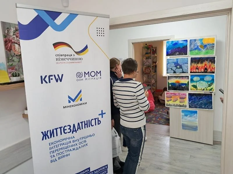 Дві жінки розглядають виставку готових живописних робіт із різнобарвним небом, закріплених на стенді. На передньому плані стоїть ролап-банер із логотипами партнерів та написом «Життєздатність+».