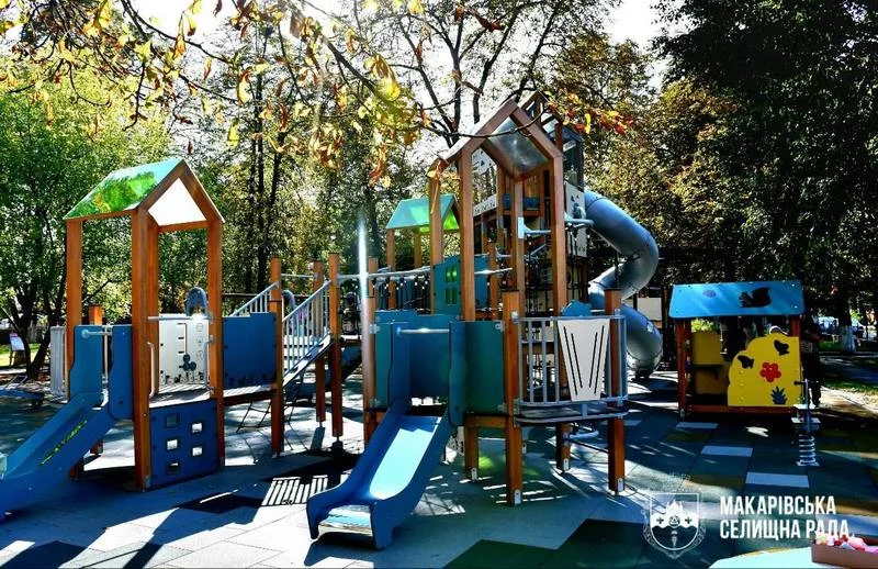 Новий сучасний дитячий майданчик у Молодіжному парку з дерев'яними ігровими вежами, гірками різної висоти, закрученою трубою-гіркою та безпечним гумовим покриттям під сонячними променями.