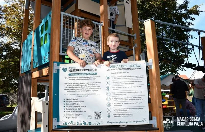 Двоє усміхнених хлопчиків стоять на ігровій конструкції поруч із білим стендом проєкту «Дитячі посмішки», на якому розміщено правила експлуатації майданчика та перелік партнерів.