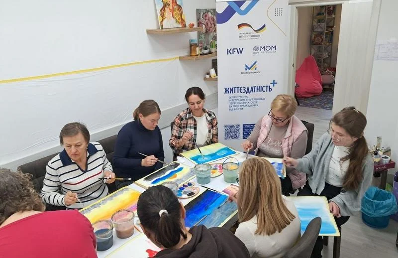 Жінки сидять за великим столом у затишному приміщенні та малюють гуашшю картини з зображенням неба і хмар. Поруч стоїть інформаційний банер проєкту «Життєздатність+».