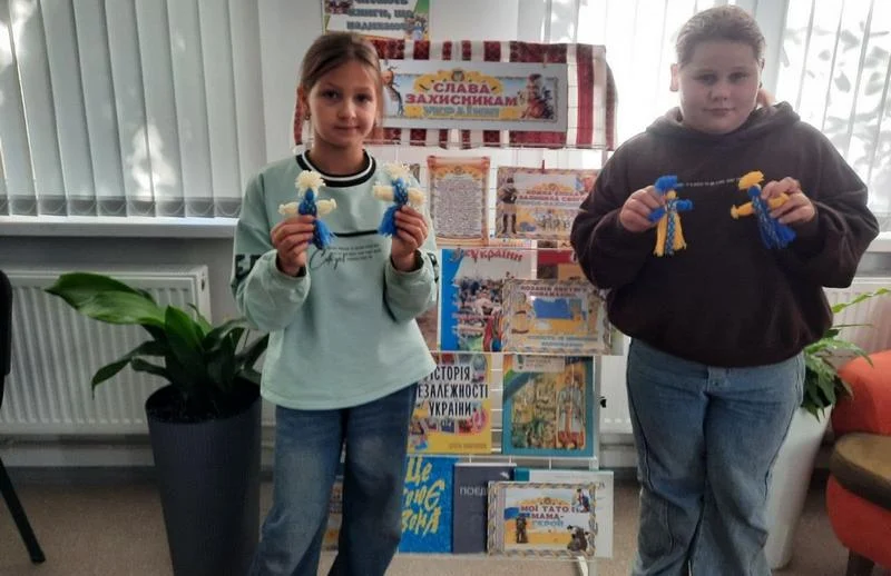 Дві дівчини-учениці тримають у руках виготовлені власноруч ляльки-мотанки із синьо-жовтих та білих ниток. На задньому плані — стенд із дитячими малюнками та книгами.