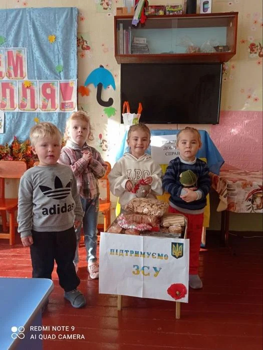 Четверо маленьких дітей у кімнаті позирують біля столика з печивом і солодощами, на якому закріплено малюнок з прапором України, маком та написом «Підтримуємо ЗСУ».