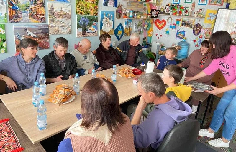 Група людей різного віку сидить навколо довгого столу, спілкуючись та пригощаючись. На столі стоять пляшки з водою та тарілки з печивом. Стіни приміщення також декоровані картинами. Дівчина в рожевій кофті роздає їжу.