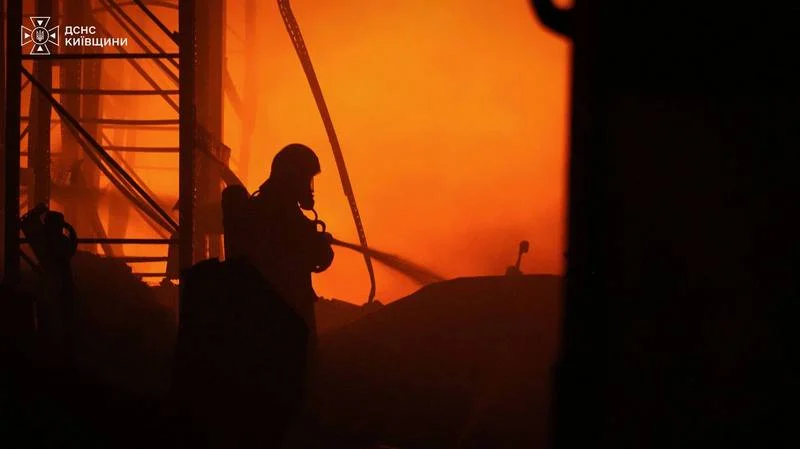 Силует рятувальника ДСНС у захисному спорядженні, який гасить полум'я на палаючому складі.