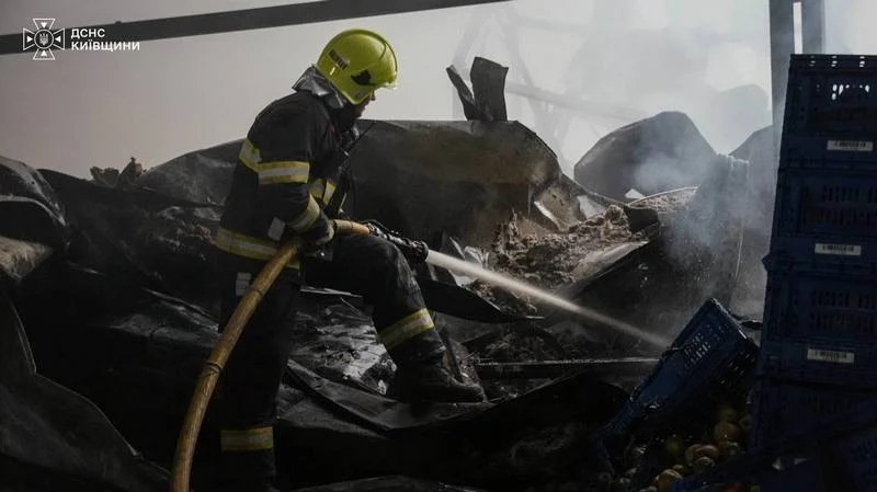 Рятувальник у захисному шоломі та формі поливає водою обвуглені конструкції складської інфраструктури.