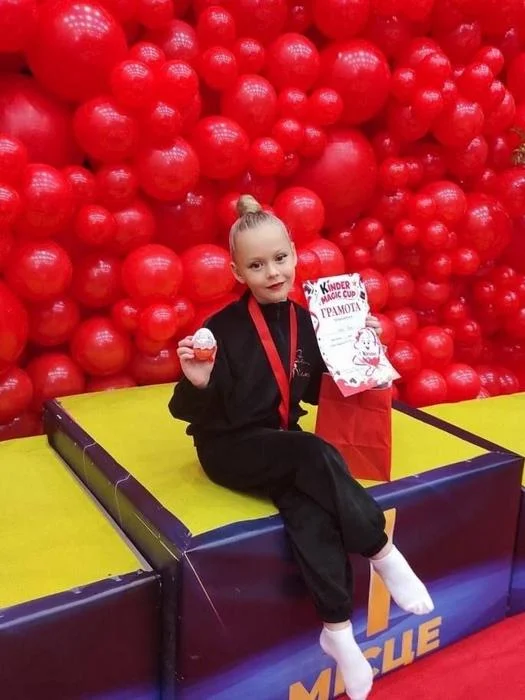 Юна призерка турніру з художньої гімнастики Kinder Magic Cup сидить на п'єдесталі пошани з медаллю на шиї, грамотою та подарунком на тлі стіни з червоних кульок.