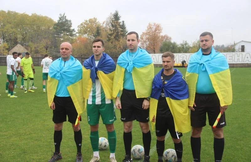 Гравці команд та арбітри шикуються на футбольному полі перед грою, накинувши на плечі державні прапори України.