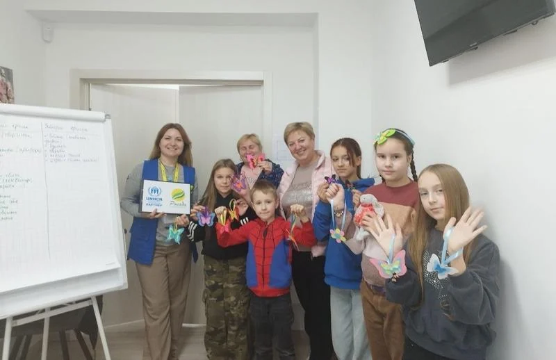 Діти та організатори стоять у кімнаті ветеранського простору та показують на камеру свої готові творчі роботи — різнобарвні фігурки метеликів, створені під час майстер-класу від БФ «Рокада».